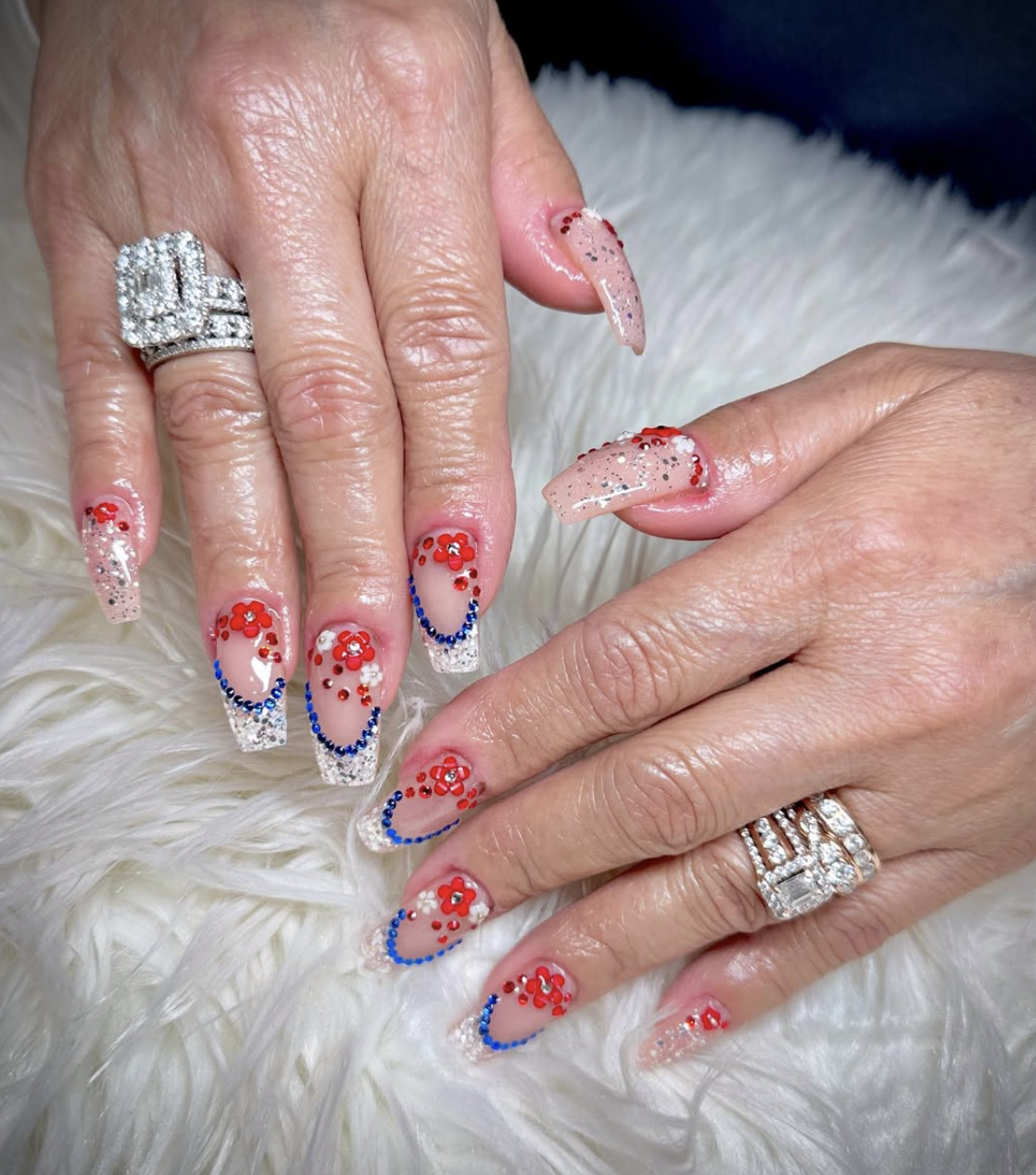 Long nude nails with crystal accents, blue curves, and red floral embellishments.